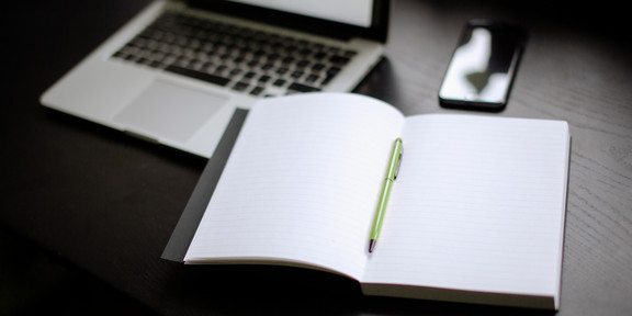 Eingeschalteter Laptop, Smartphone und Notizbuch mit Stift auf einem Tisch liegend.